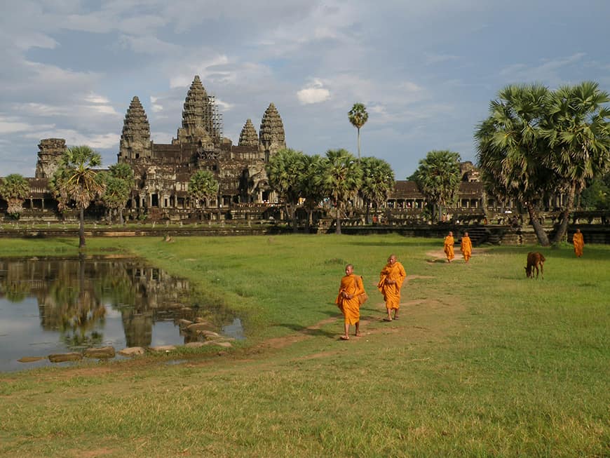 Grupa buddyjskich mnichów idąca ścieżką przez trawnik przed Angkor Wat. Monumentalna świątynia i jej wieże są widoczne w tle, a część kompleksu odbija się w wodzie.